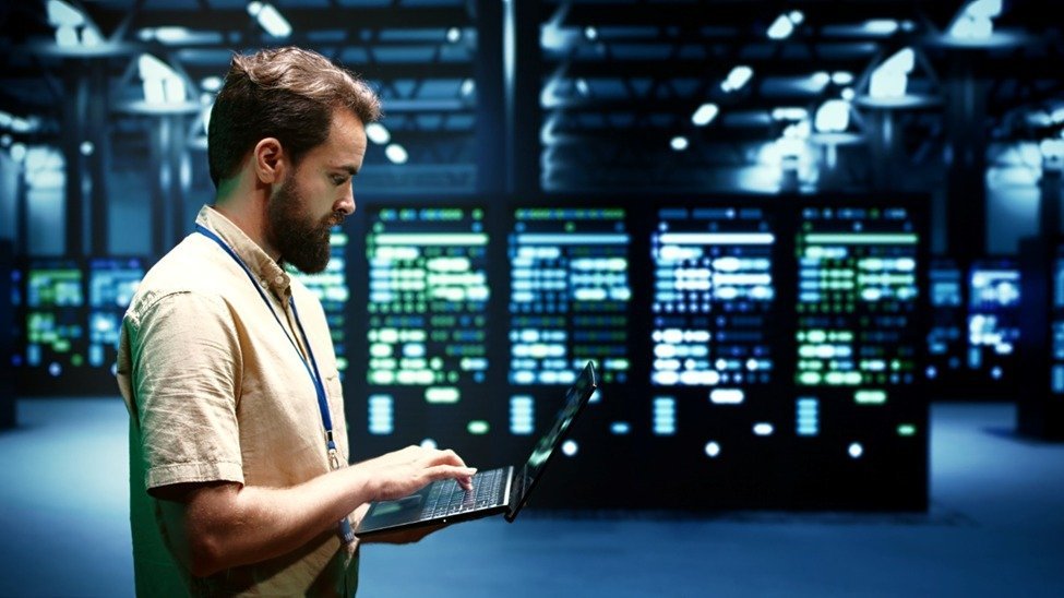 Man with a beard typing on a laptop in a server room with illuminated data panels.